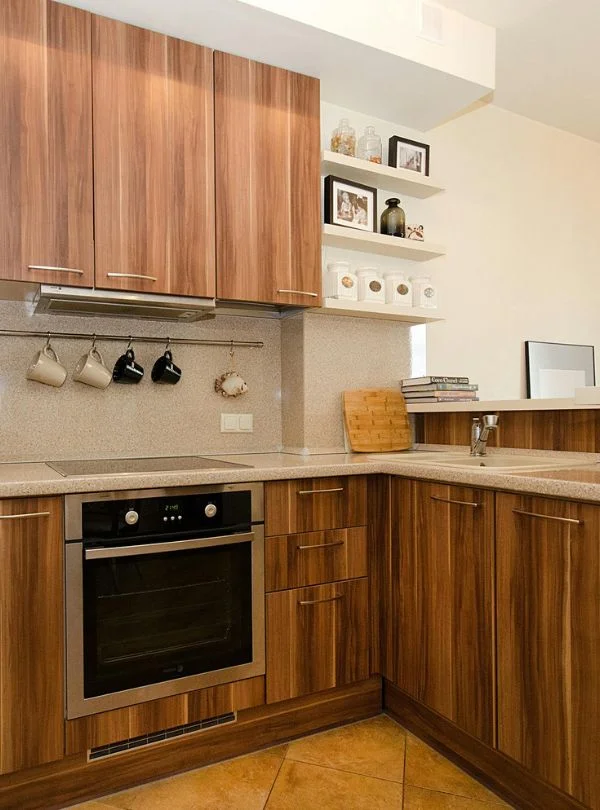Cocina en L con muebles de madera y encimeras claras, diseño funcional y acogedor con horno empotrado y estanterías decorativas